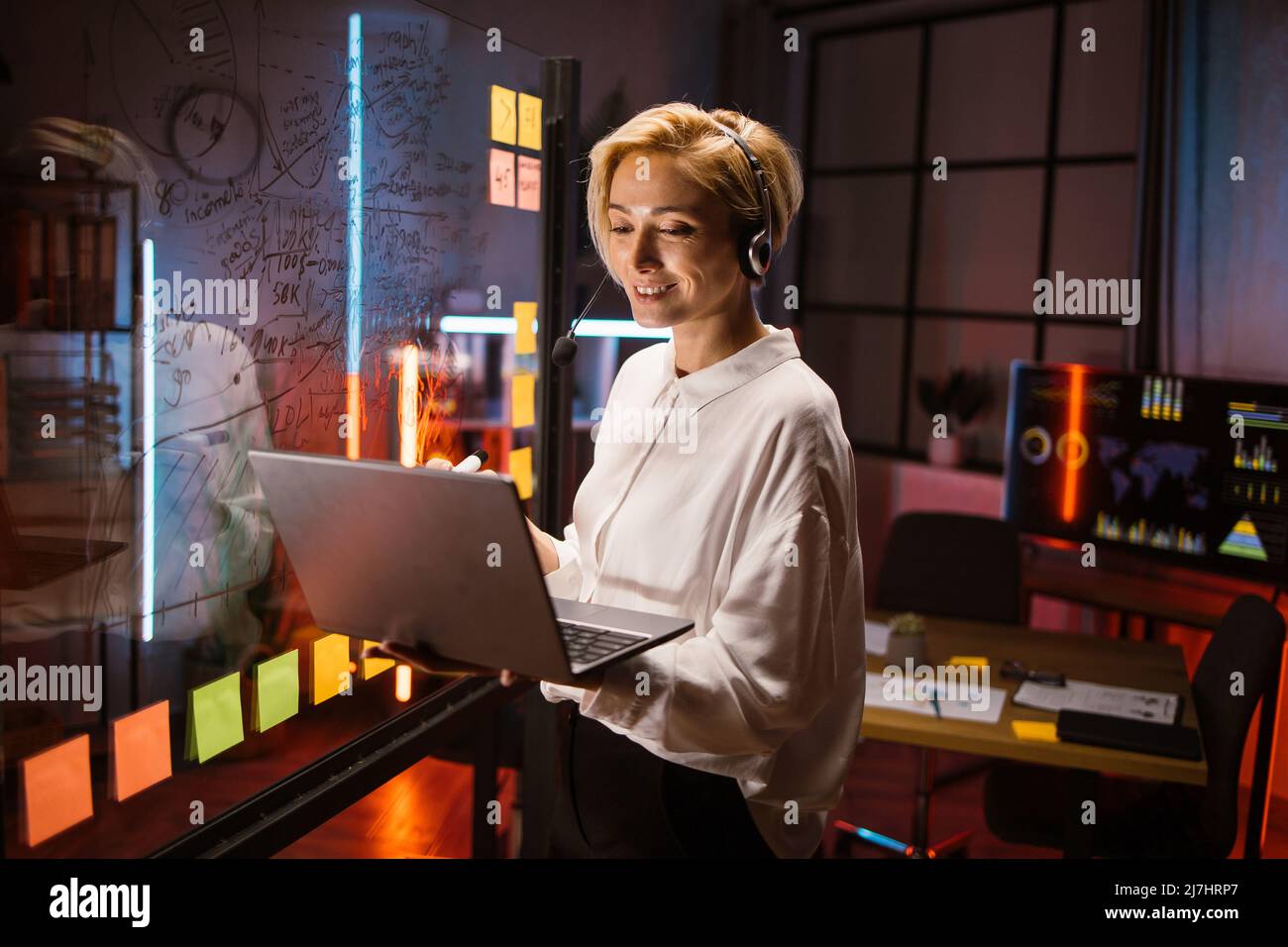 Smiling Caucasian businesswoman in headset, using laptop for remote brainstorm meeting with colleagues, standing near glass board with colorful sticky paper notes Stock Photo