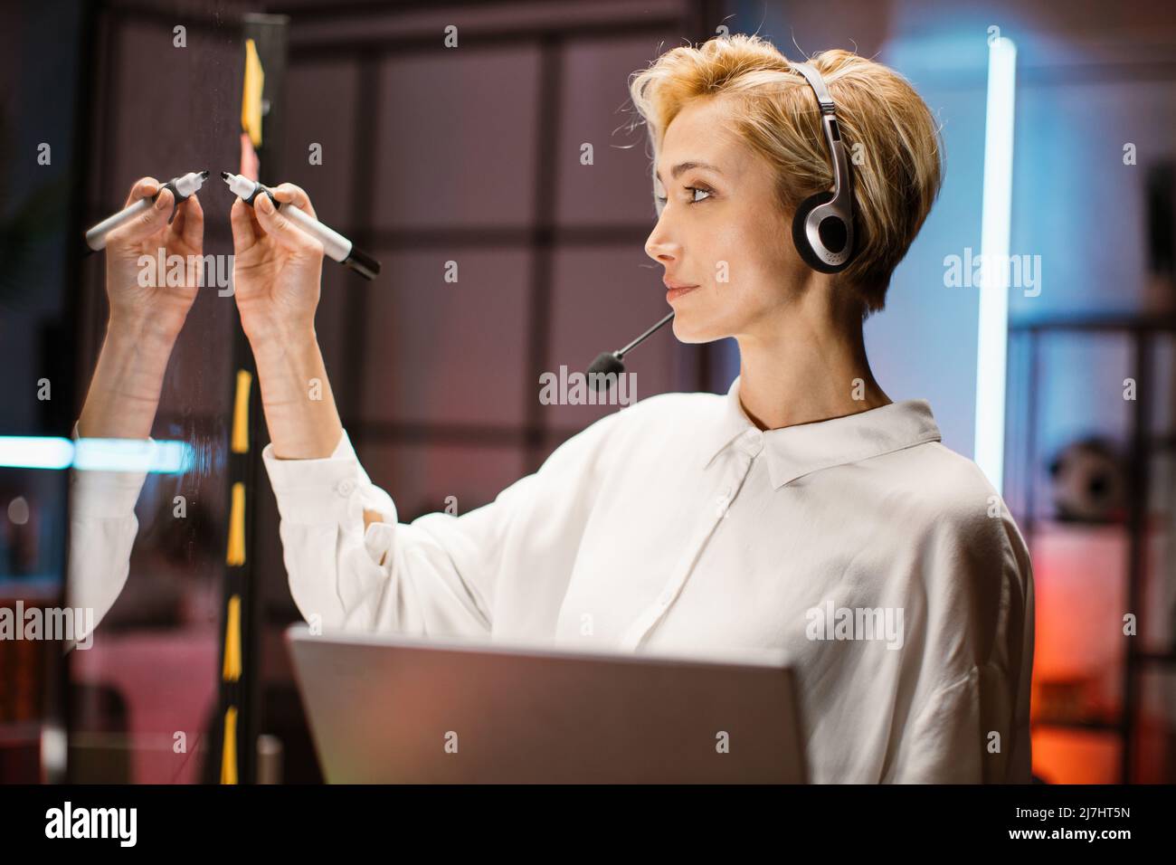 Close up side view of confident blond young businesswoman wearing white shirt and headset, writing down plans and ideas on a glass office panel while holding in her hands laptop Stock Photo