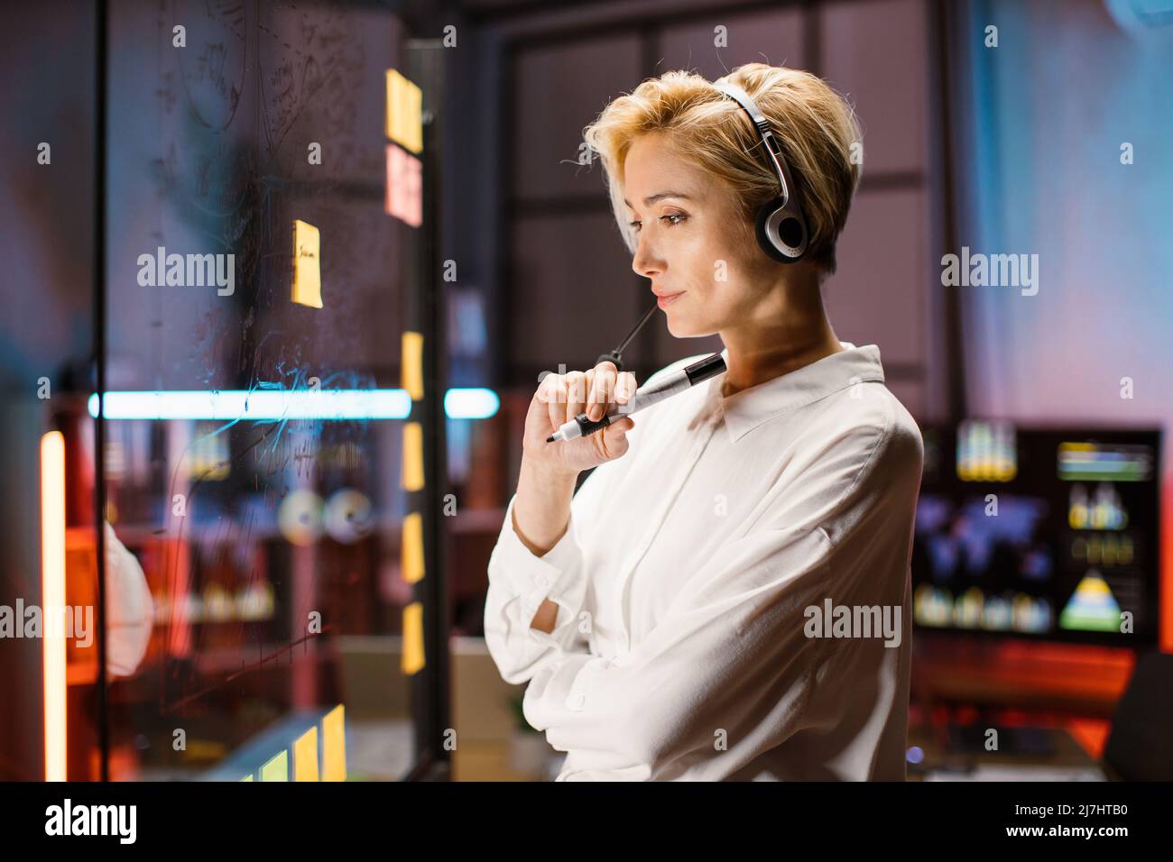 Pensive smart blond businesslady in headset, holding marker in hands and looking at the transparent glass board, while working late at dark office. Business, brainstorming, planning concept Stock Photo