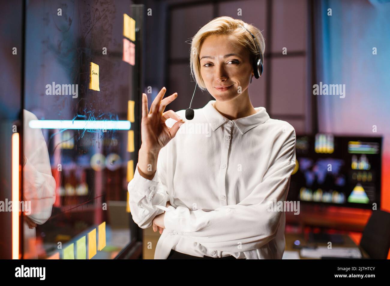 Confident Caucasian female business leader in white shirt and headset, posing near transparent glass board with marker pen, looking at camera and smiling. Project deadline, work planning concept Stock Photo