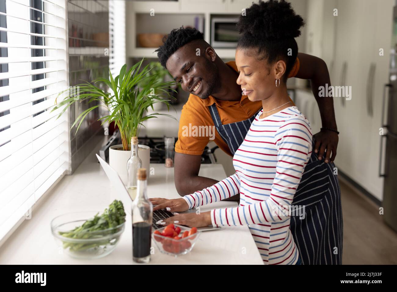 Smiling african american young man looking at girlfriend using laptop on kitchen counter at home Stock Photo