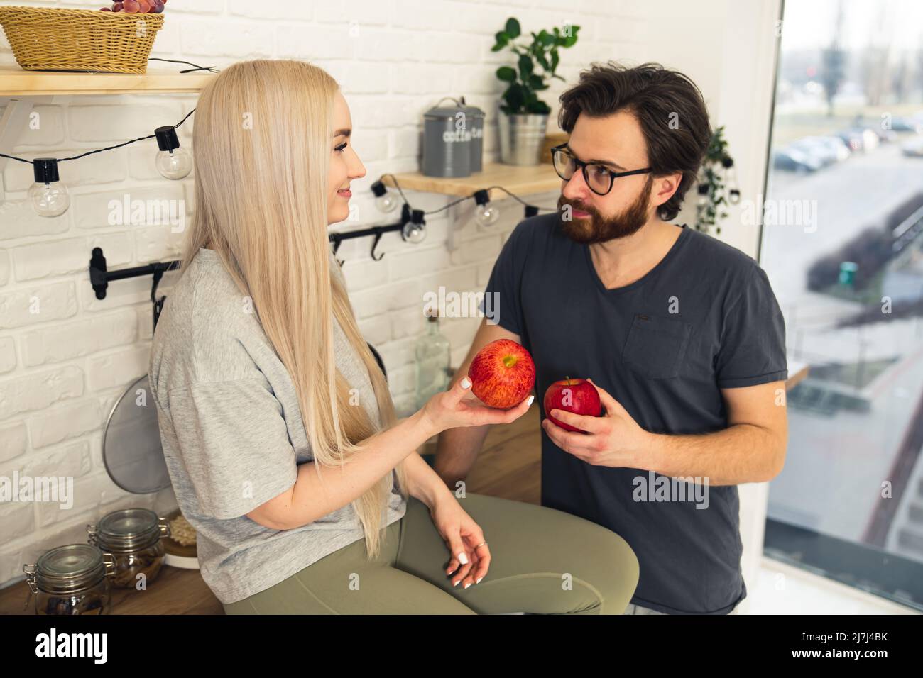 Togheterness concept. Caucasian woman and man in their 20s looking at each other with love and affection holding red juicy apples. Woman sitting on a kitchen counter, man standing near her. High quality photo Stock Photo