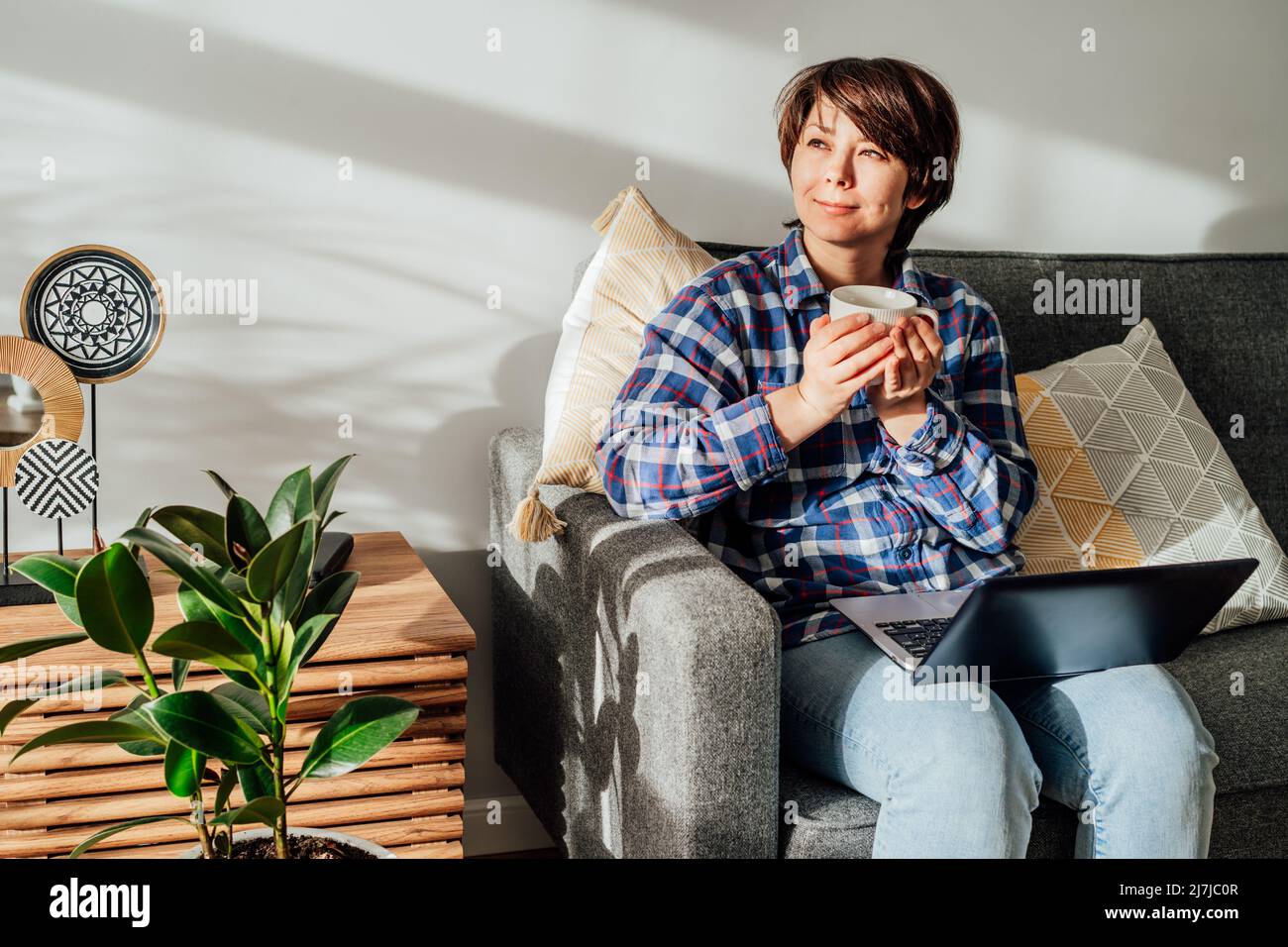 Dreaming woman using laptop and drinking coffee, tea at home sitting on sofa, couch in sunlight beams. Cozy workspace in modern interior with green pl Stock Photo