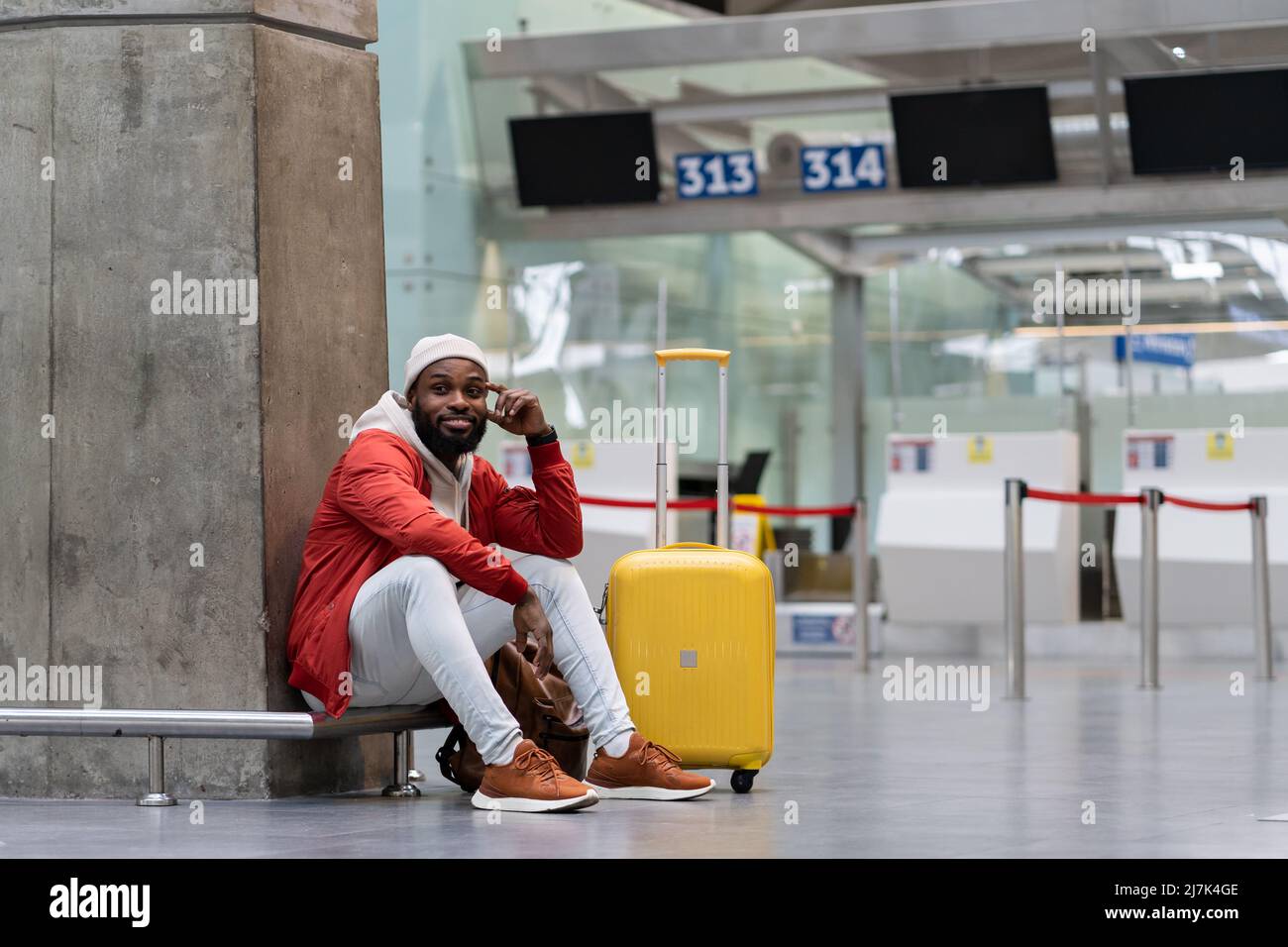 Joyful African American man sitting in airport terminal with yellow suitcase near check-in counters. Stock Photo