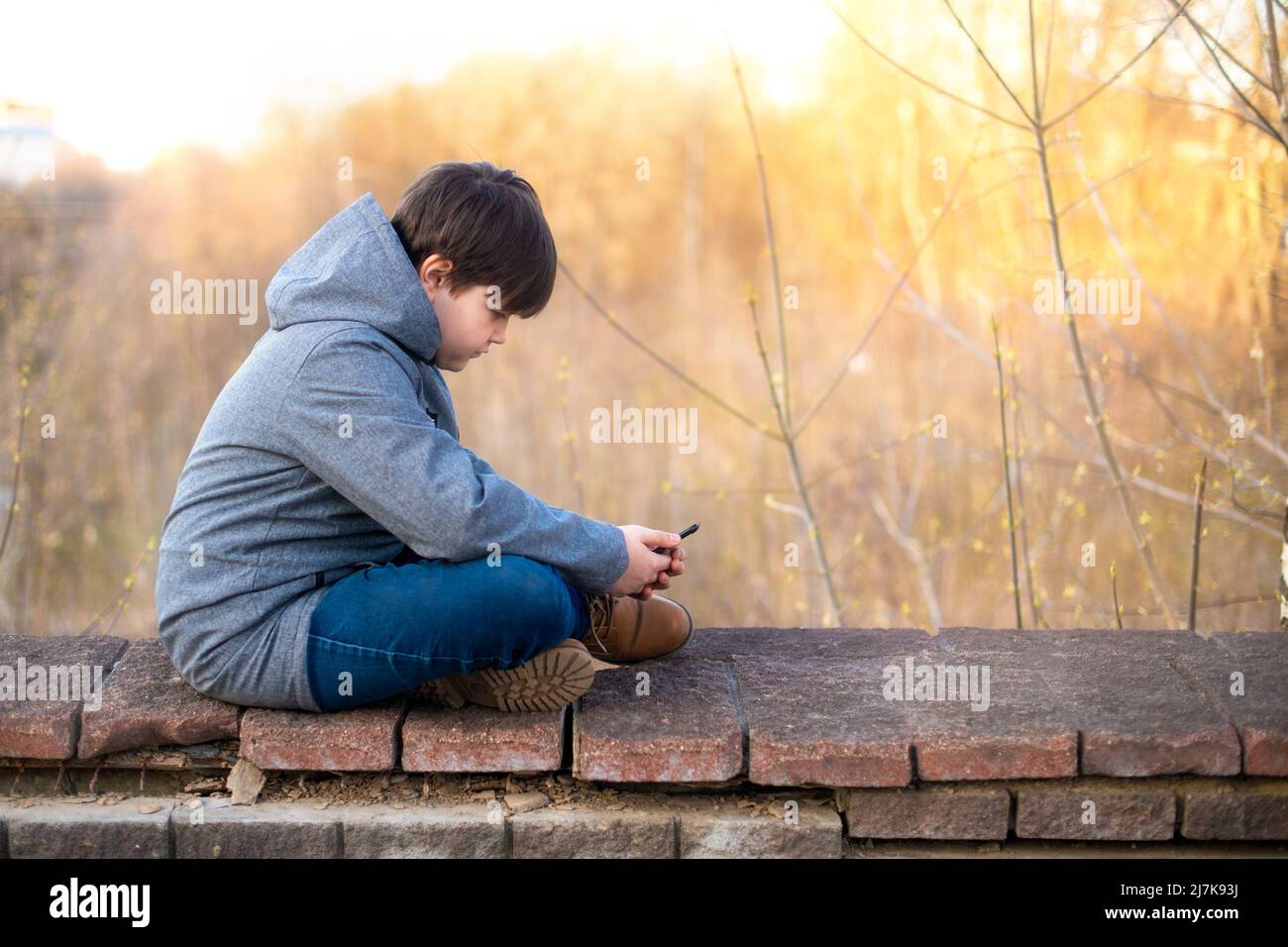 A schoolboy boy uses a mobile phone in his hands in nature in the city in the spring at sunset. Free time. Stock Photo