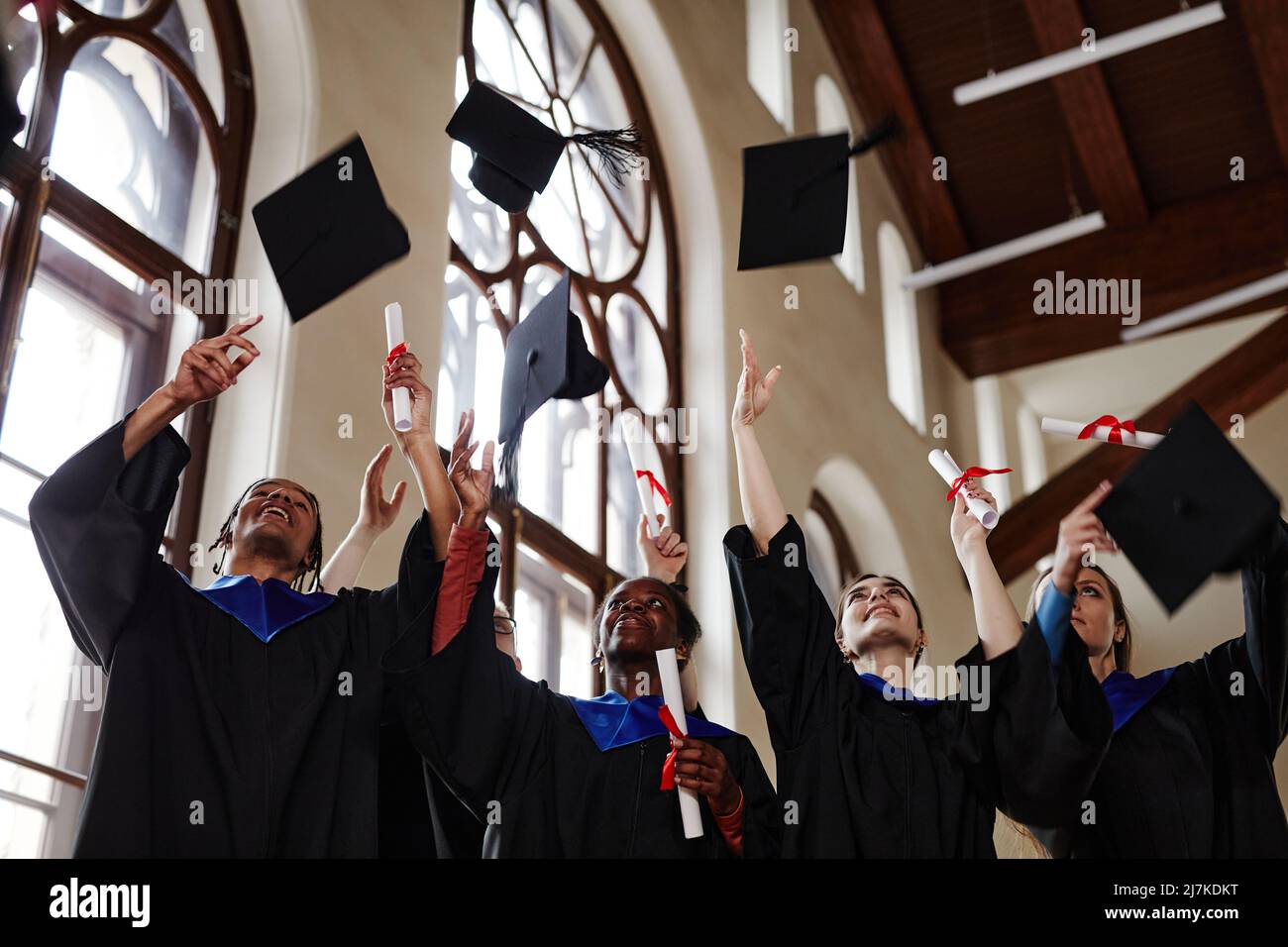 Group of joyful young people wearing graduation gowns at ceremony in university and throwing caps in air Stock Photo