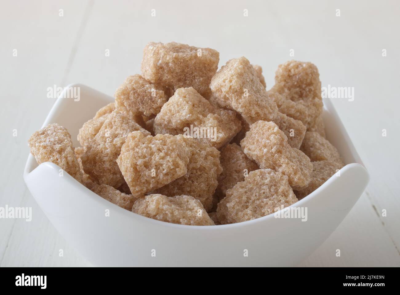 Cube-shaped cane sugar in porcelain bowl on white wooden table. Front closeup view, no people. Stock Photo