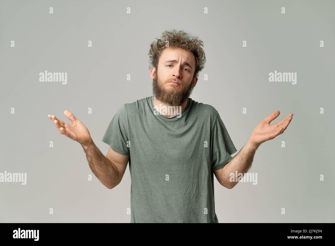 Gesturing I DONT KNOW or I AM SORRY young handsome bearded wild curly hair man with bright blue eyes isolated on grey background. Young thinking man in green t shirt on white. Copy space.  Stock Photo