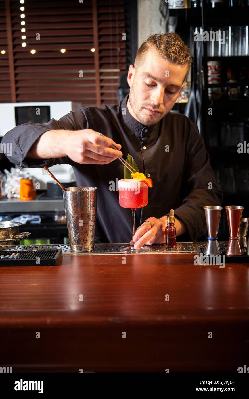 Stylish young male barkeeper with braided hair in black uniform decorating delicious pink cocktail with fresh leaf during work in bar Stock Photo
