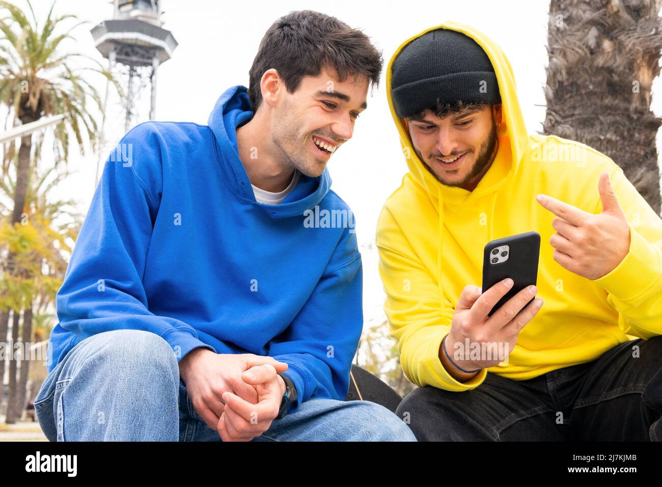 Two cheerful friends with colorful outfits using phone while sitting on street Stock Photo