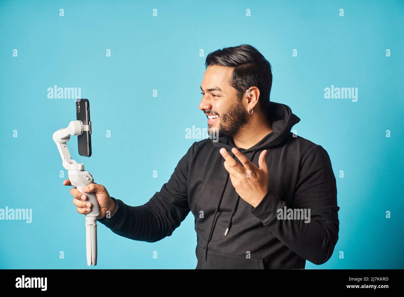 Positive bearded male blogger in black hoodie recording vlog on modern smartphone while standing against blue background in light studio Stock Photo