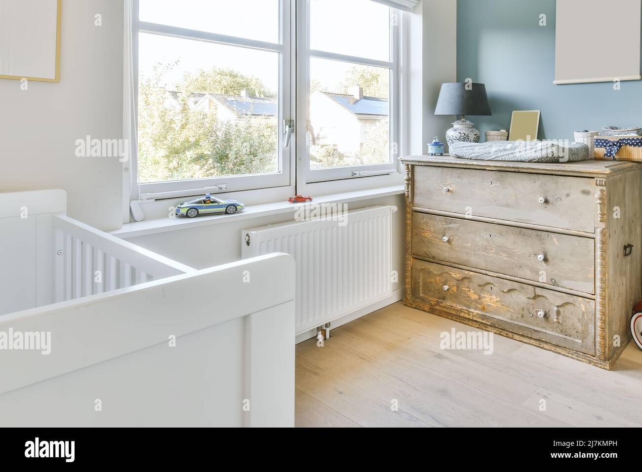 White crib placed at wall near window in light spacious children bedroom with wooden chest of drawers with lamp at home Stock Photo