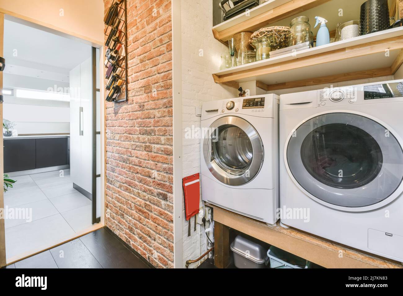 Modern white electric washing machines placed on shelf near various utensils in light laundry room with brick wall and opened room Stock Photo