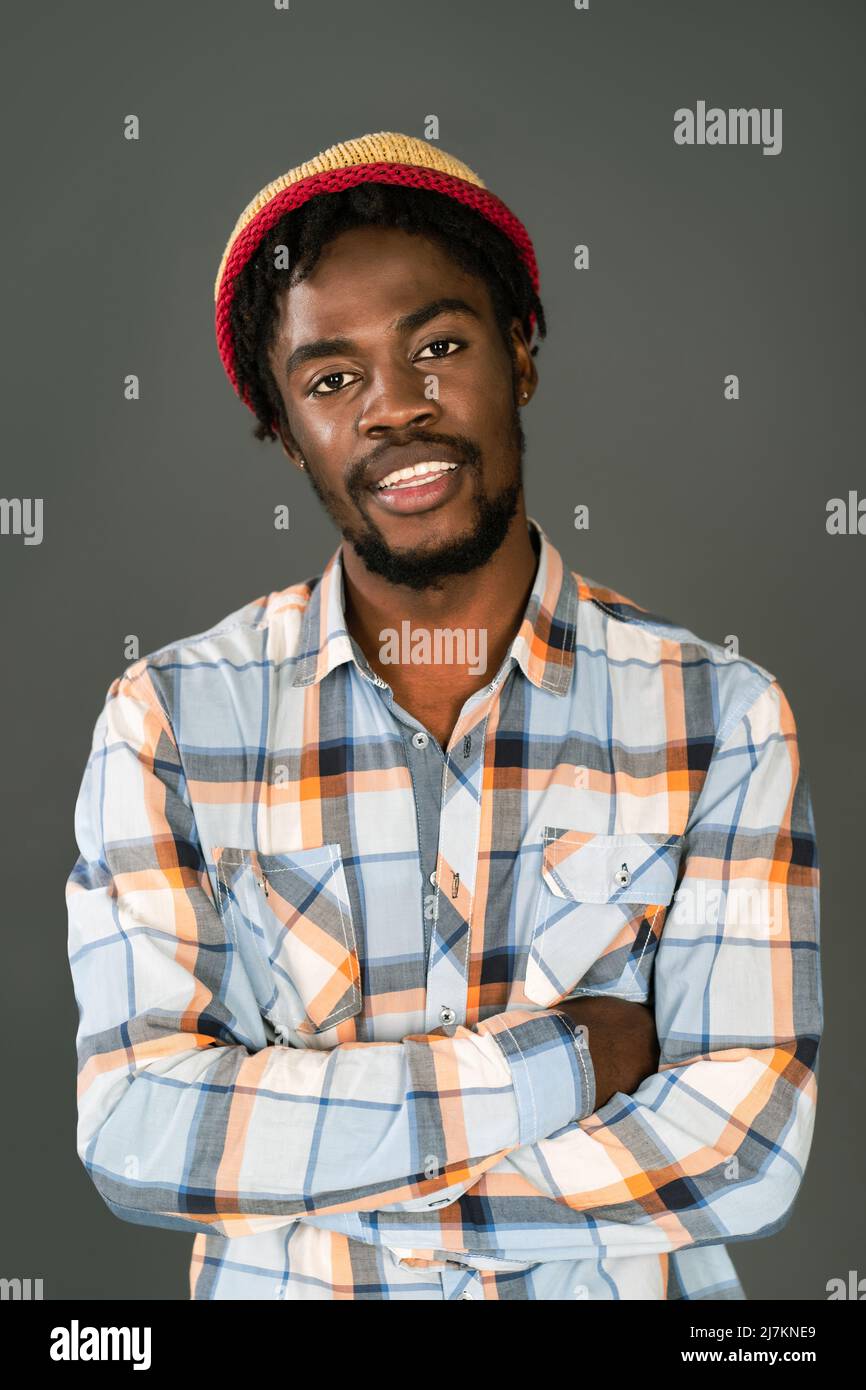 Young smiling black man in afro hat standing with arms folded looking at camera. Cheerful african american young man in plaid shirt looking at camera isolated on gray background.  Stock Photo
