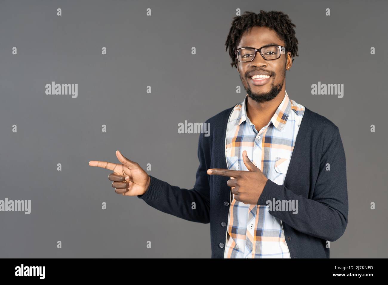 Smiling black young man in glasses pointing with hands sideways looking at camera. Cheerful african american young man in plaid shirt looking at camera isolated on gray background.  Stock Photo
