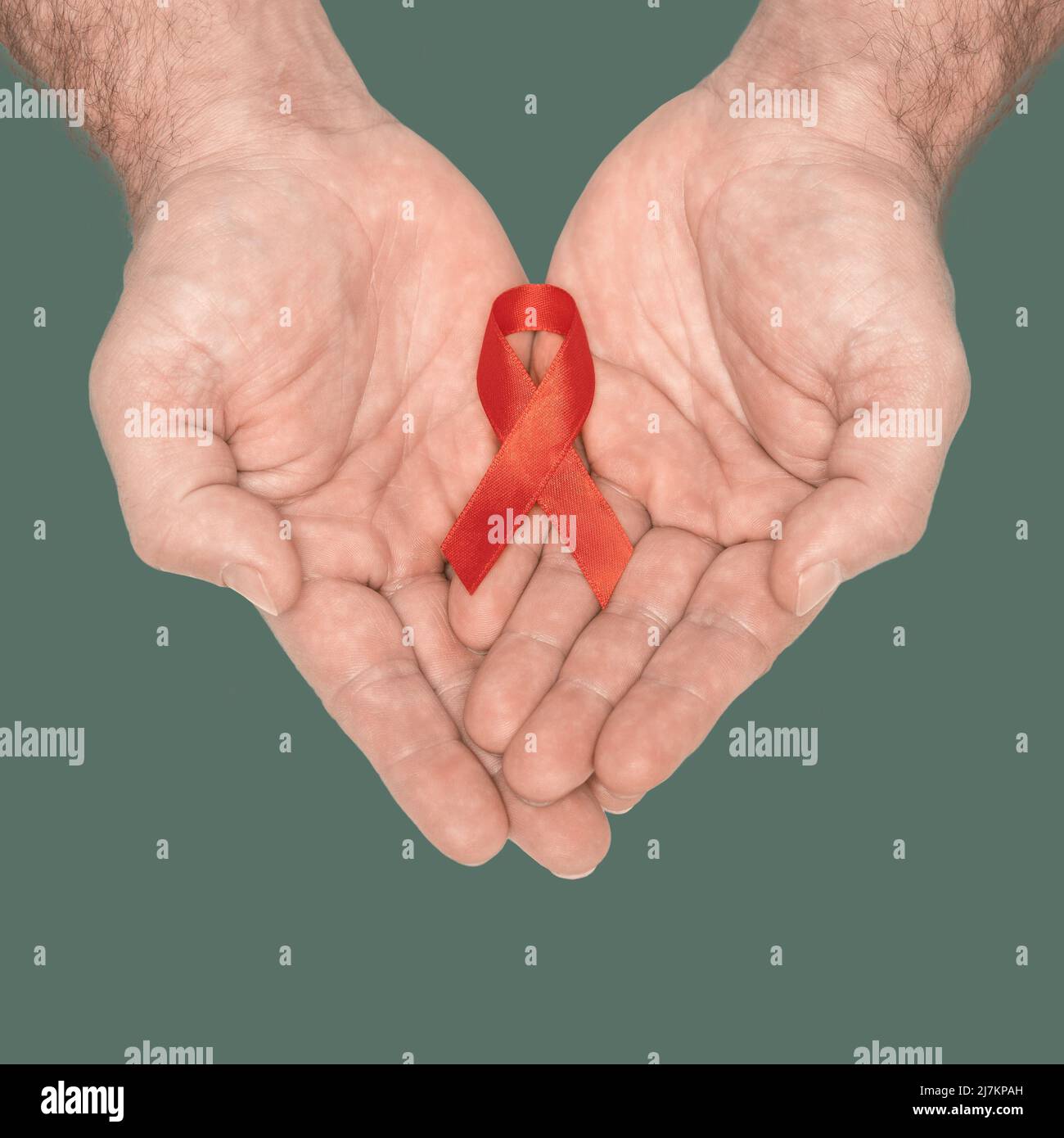 Red awareness ribbon bow on mans helping hands isolated on green background. HIV, AIDS world day. Social life issues concept. Aids charity fund concept. Healthcare and medicine concept.  Stock Photo