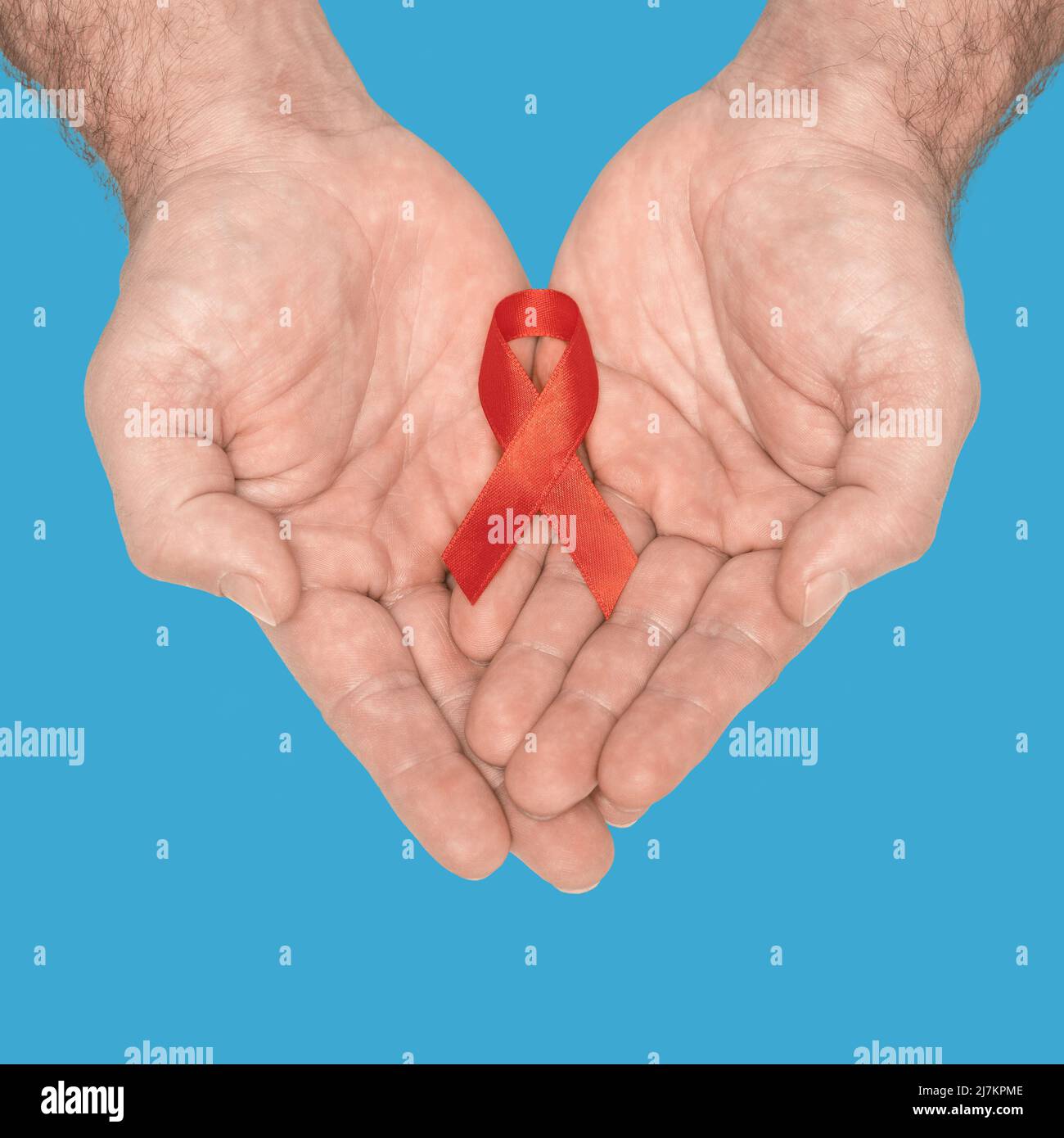 Red awareness ribbon bow on mans helping hands isolated on blue background. HIV, AIDS world day. Social life issues concept. Aids charity fund concept. Healthcare and medicine concept.  Stock Photo