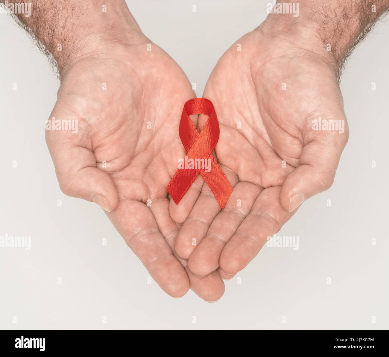 Red awareness ribbon bow on mans helping hands isolated on white background. HIV, AIDS world day. Social life issues concept. Healthcare and medicine concept. Aids charity fund concept.  Stock Photo