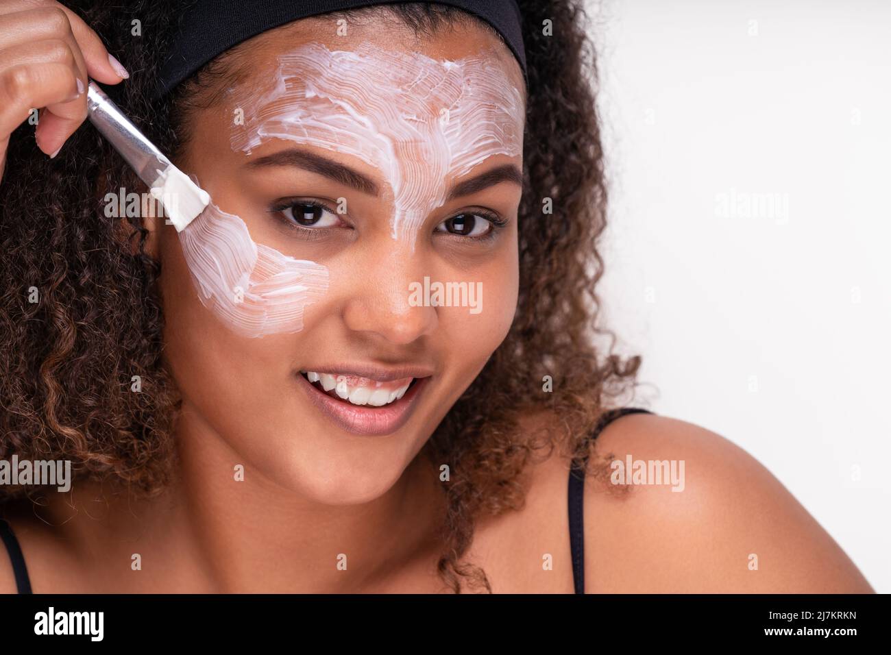 Self assured smiling young ethnic female model with curly dark hair and in headband applying mask on face with brush and looking at camera against whi Stock Photo