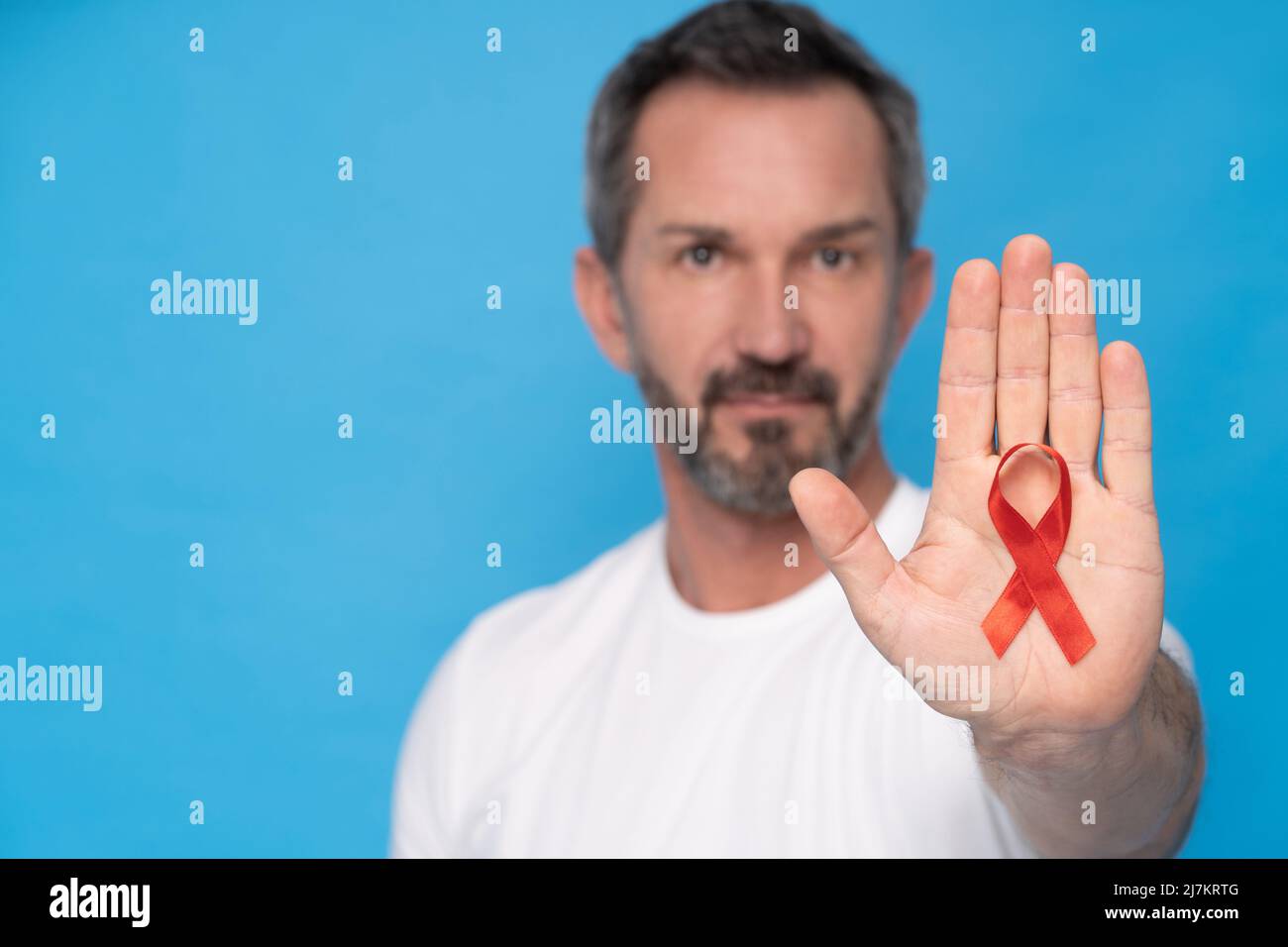 Man hold red ribbon bow AIDS awareness symbol on a heart wearing a white t-shirt isolated on a blue background. Modern medicine and healthcare. AIDS awareness concept. Stock Photo
