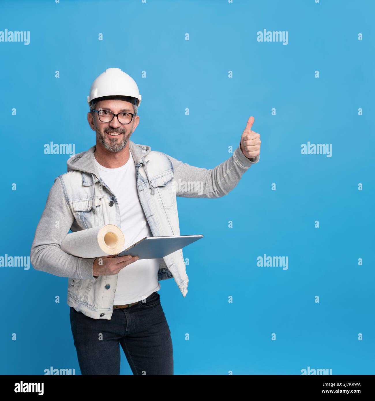 Middle aged handsome architecture man holding roll of wall paper and digital tablet pc in trendy casual denim outfit, hard hat or helmet gesturing thumb up or perfect isolated on blue background.  Stock Photo