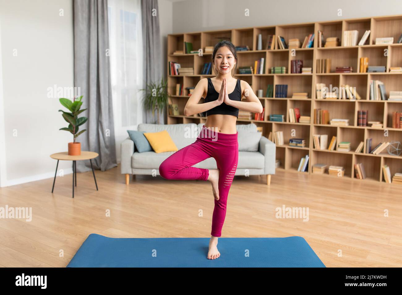 Full length shot of calm asian lady in sports clothes standing in tree pose, practicing yoga on mat at home Stock Photo