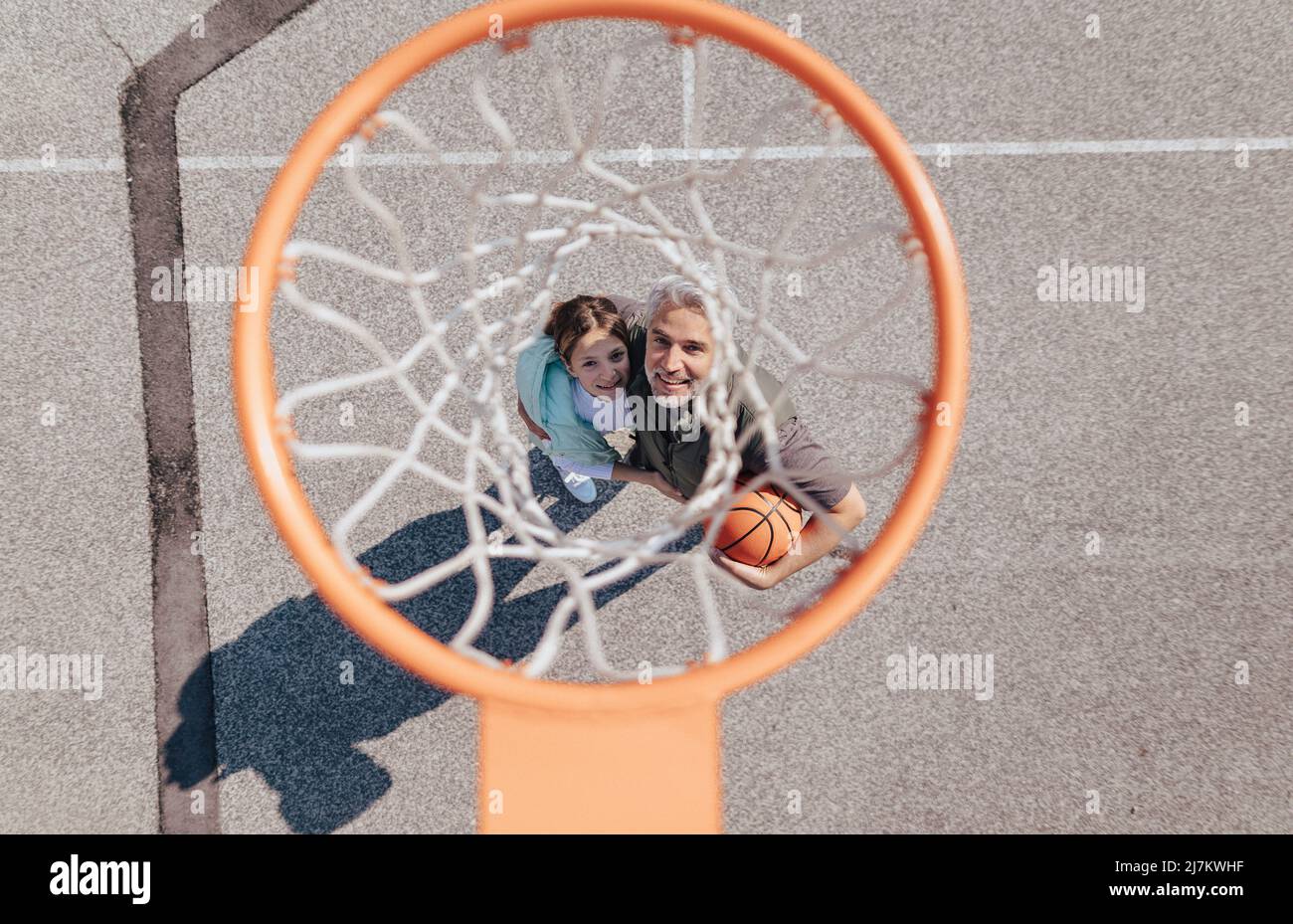 Happy father and teen daughter embracing and standing under a basketball hoop net, directly above Stock Photo