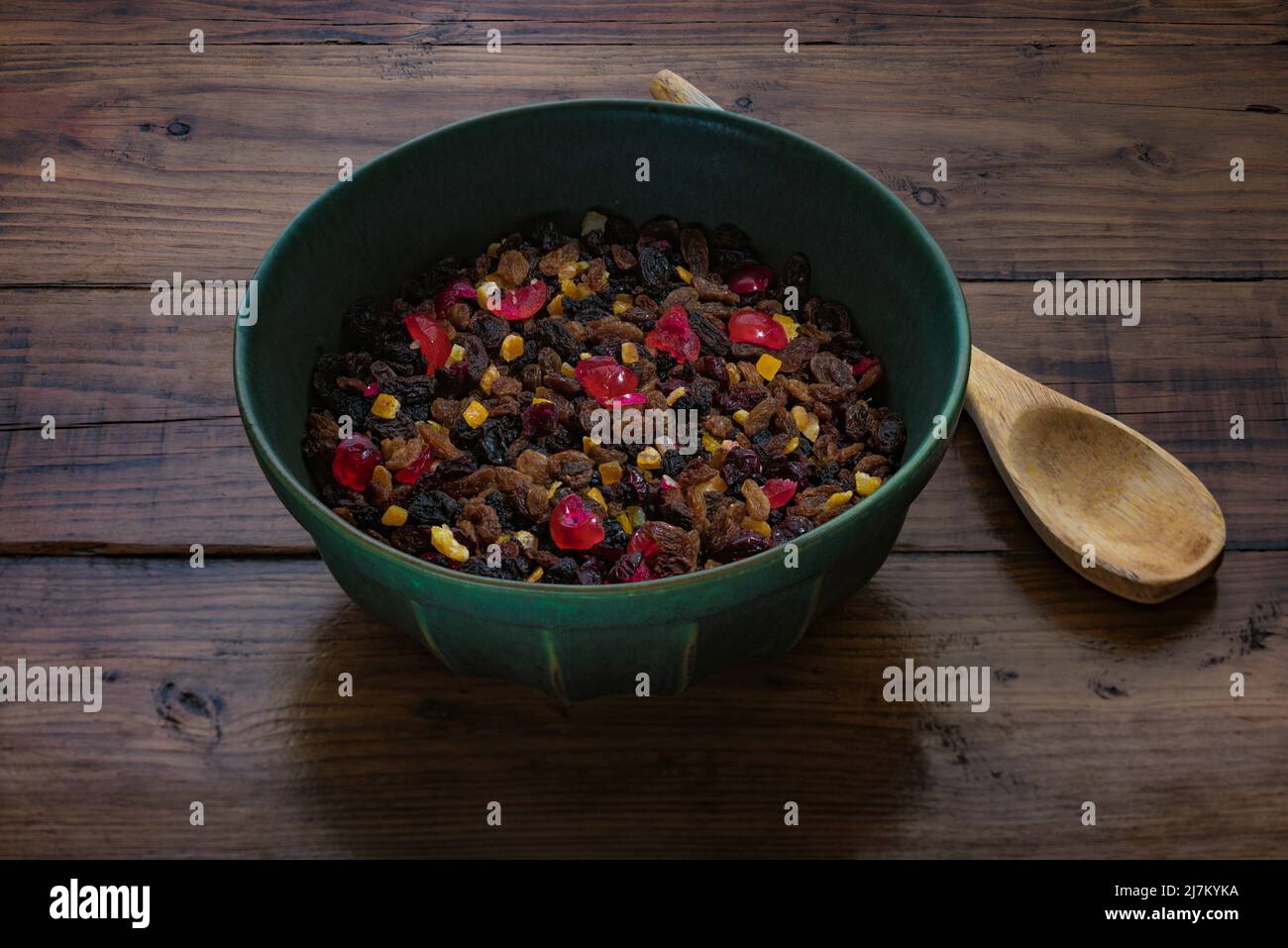 Dark green mixing bowl containing mixed dried fruit, along with a wooden spoon on a dark wooden work surface. Stock Photo
