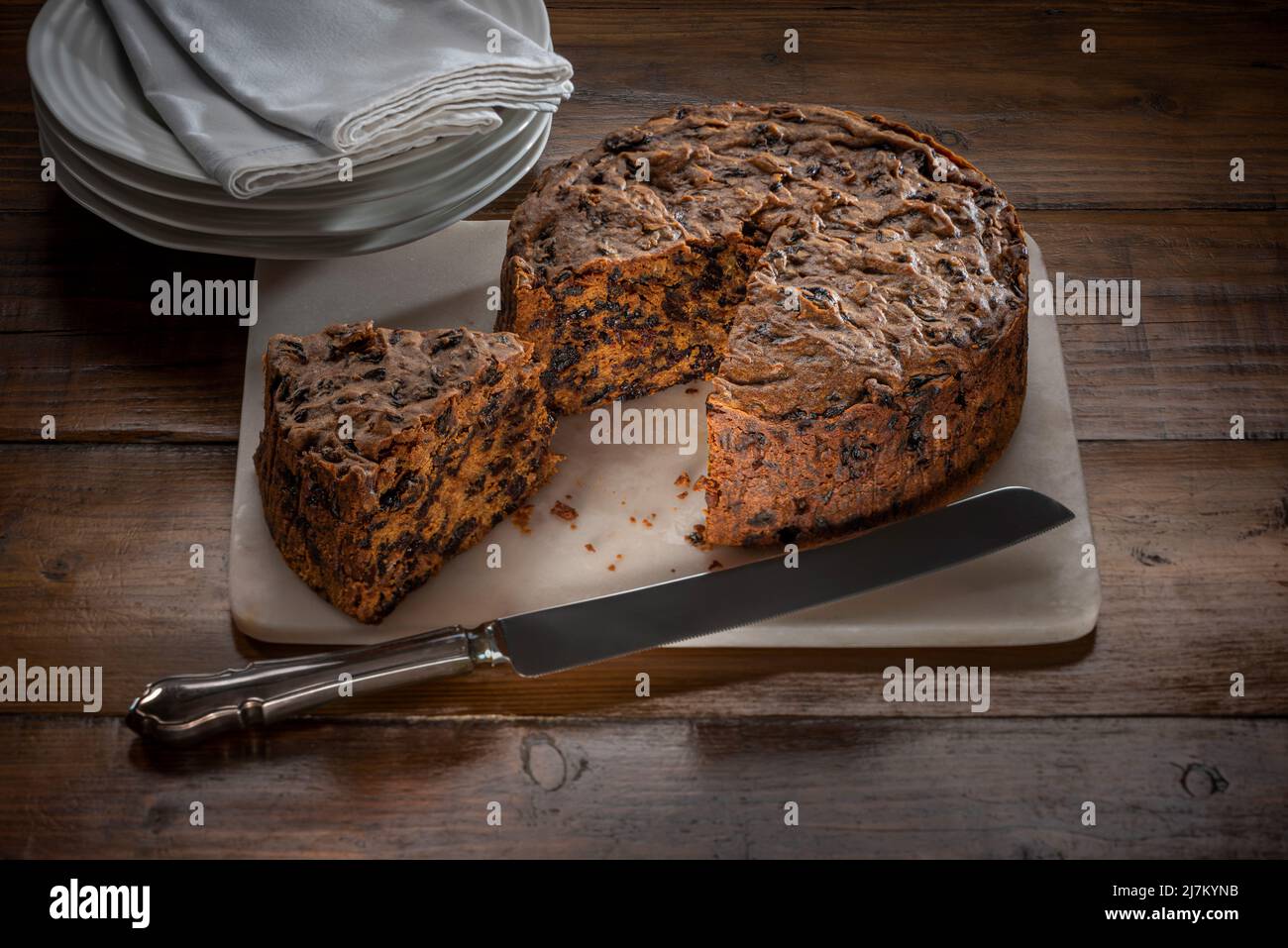 Fruit cake served on a marble board with a segment cut out, on a dark wood work top, with a stack of white plates and napkins in the background. Stock Photo