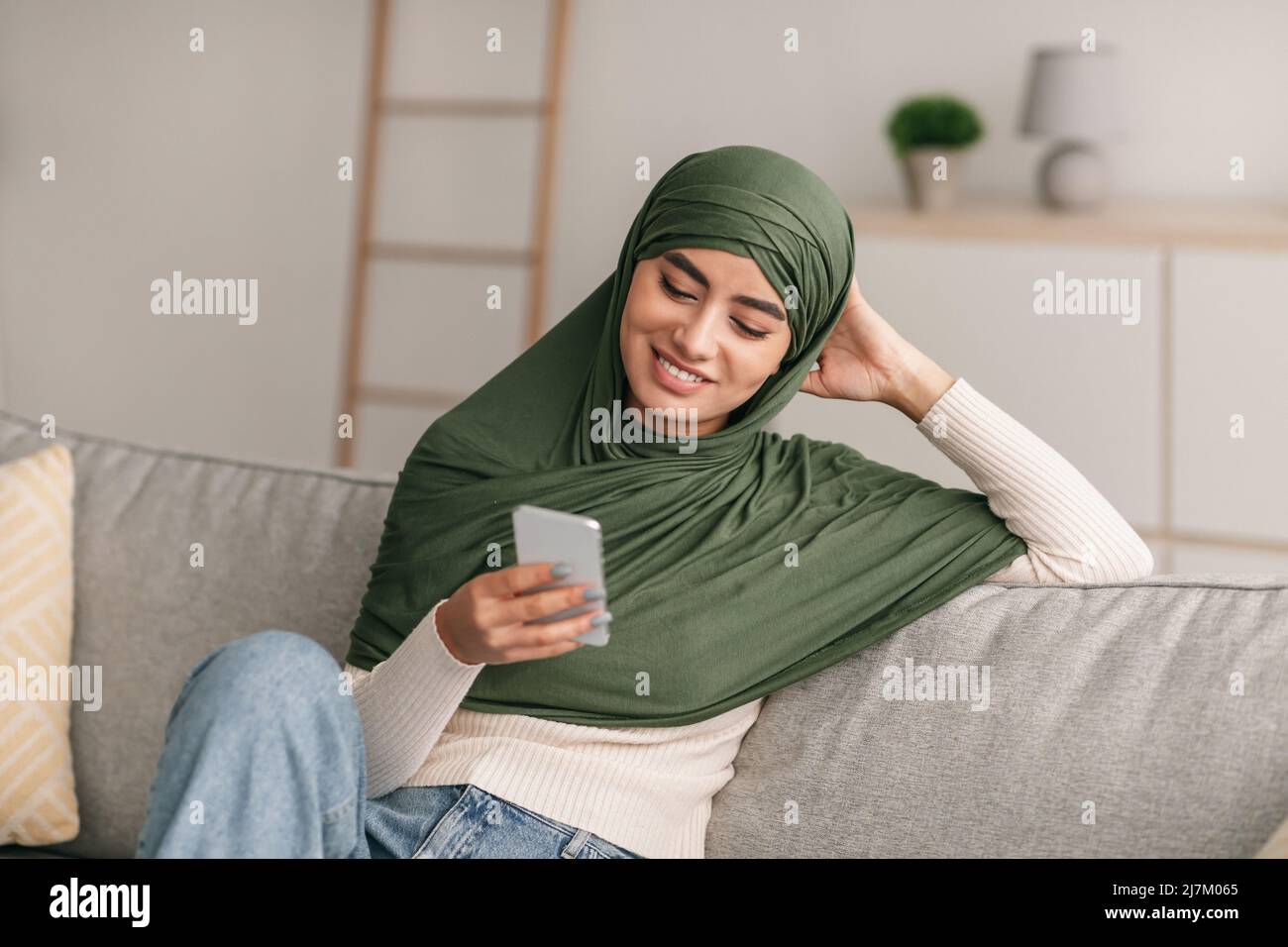Positive young Arab lady in hijab sitting on couch, having video call ...