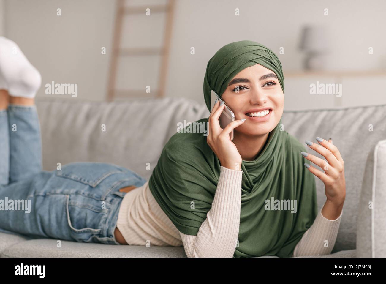 Positive young Arab lady in hijab lying on sofa, making call on mobile ...