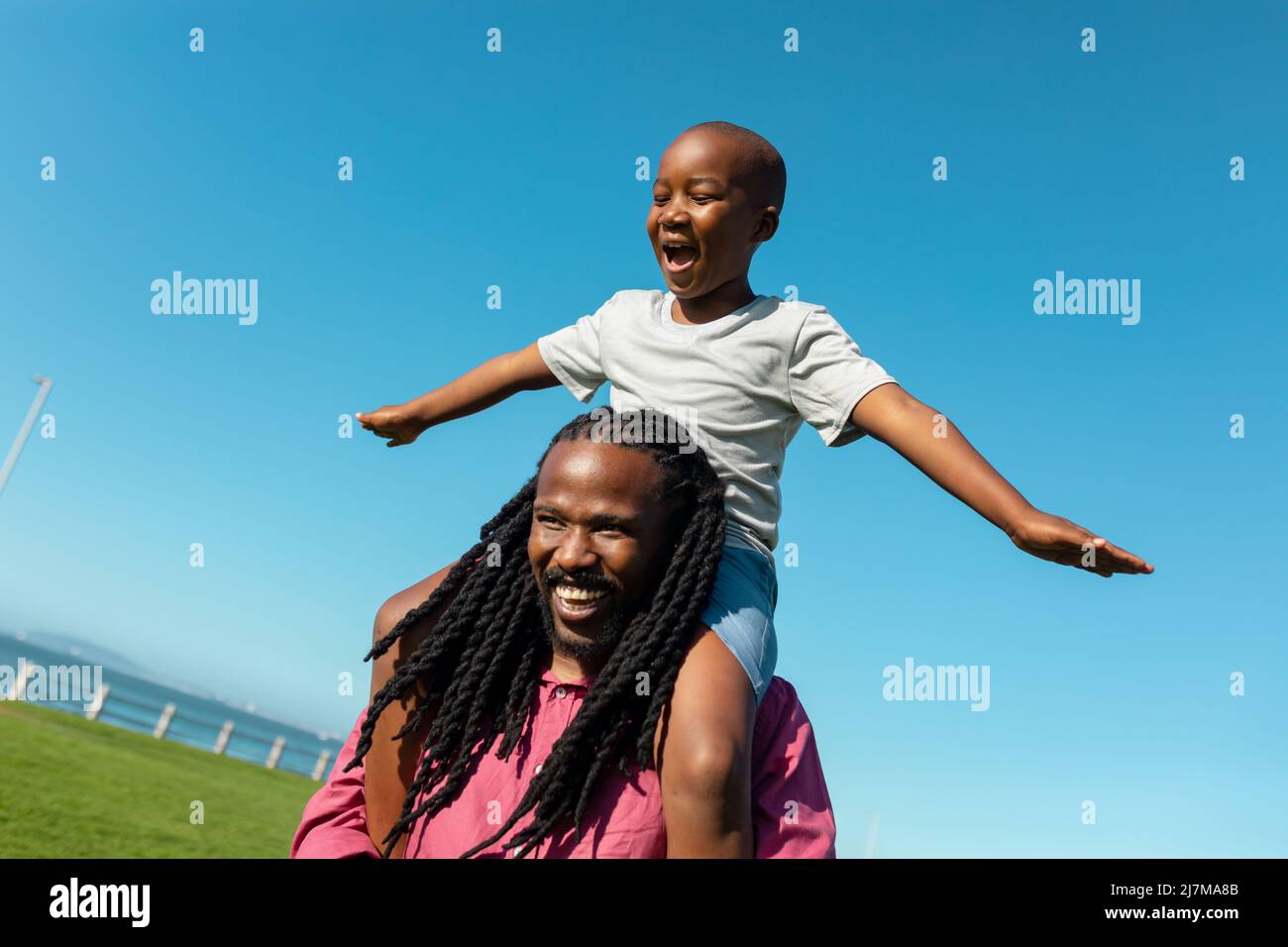 African american father carrying son with arms outstretched on shoulders against clear blue sky Stock Photo