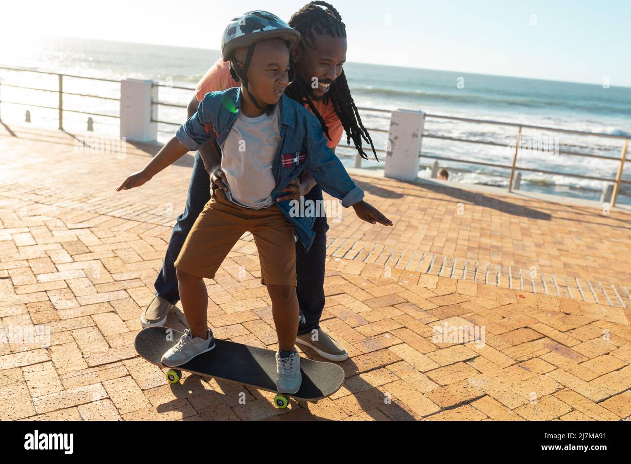 African american father support son learning skateboarding on promenade during sunny day Stock Photo