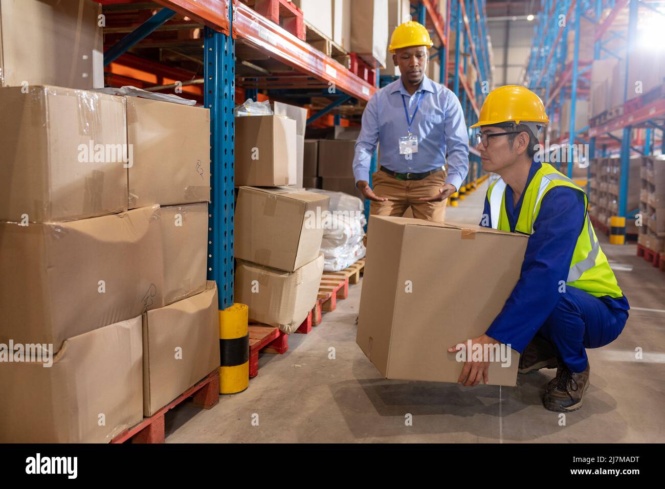 African american young foreman guiding asian mature male worker while lifting cardboard box by shelf Stock Photo