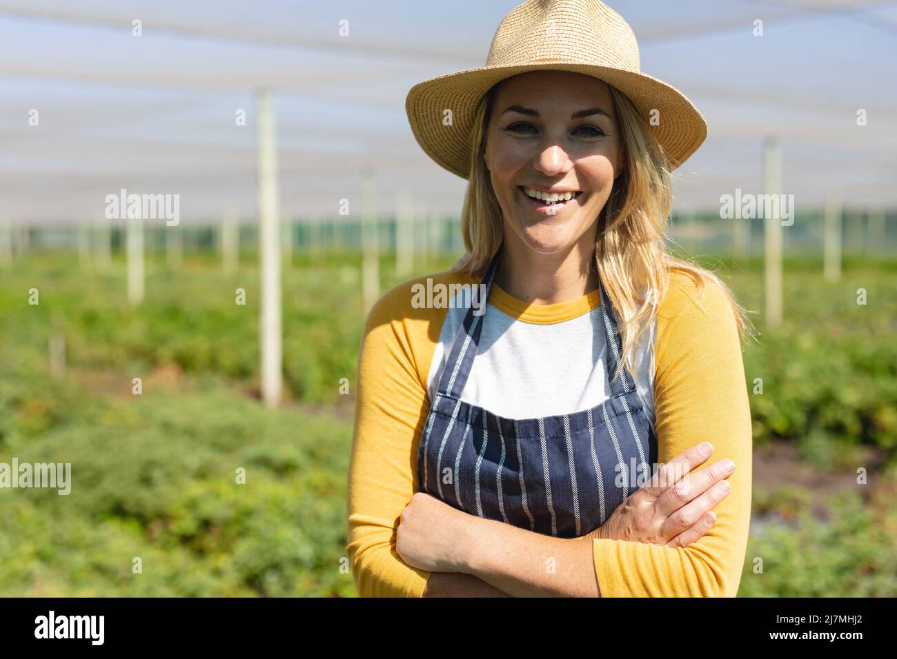 Portrait of smiling caucasian mid adult female farmer wearing hat with arms crossed in greenhouse Stock Photo