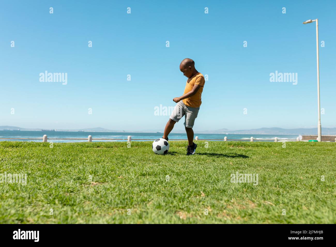 Full length of african american boy kicking soccer ball on grass against blue sky with copy space Stock Photo