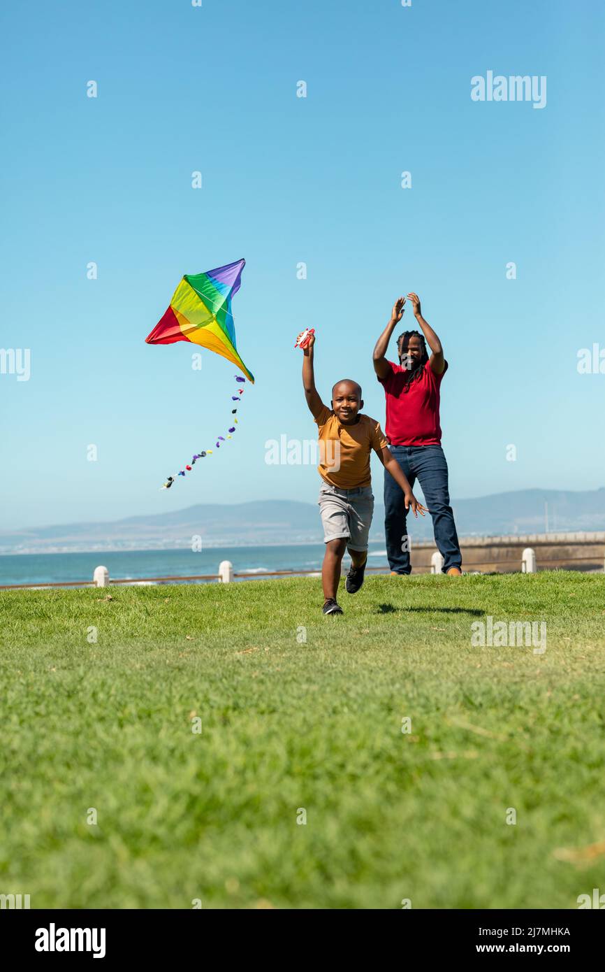 African american boy running with kite while father cheering on grassy promenade against blue sky Stock Photo