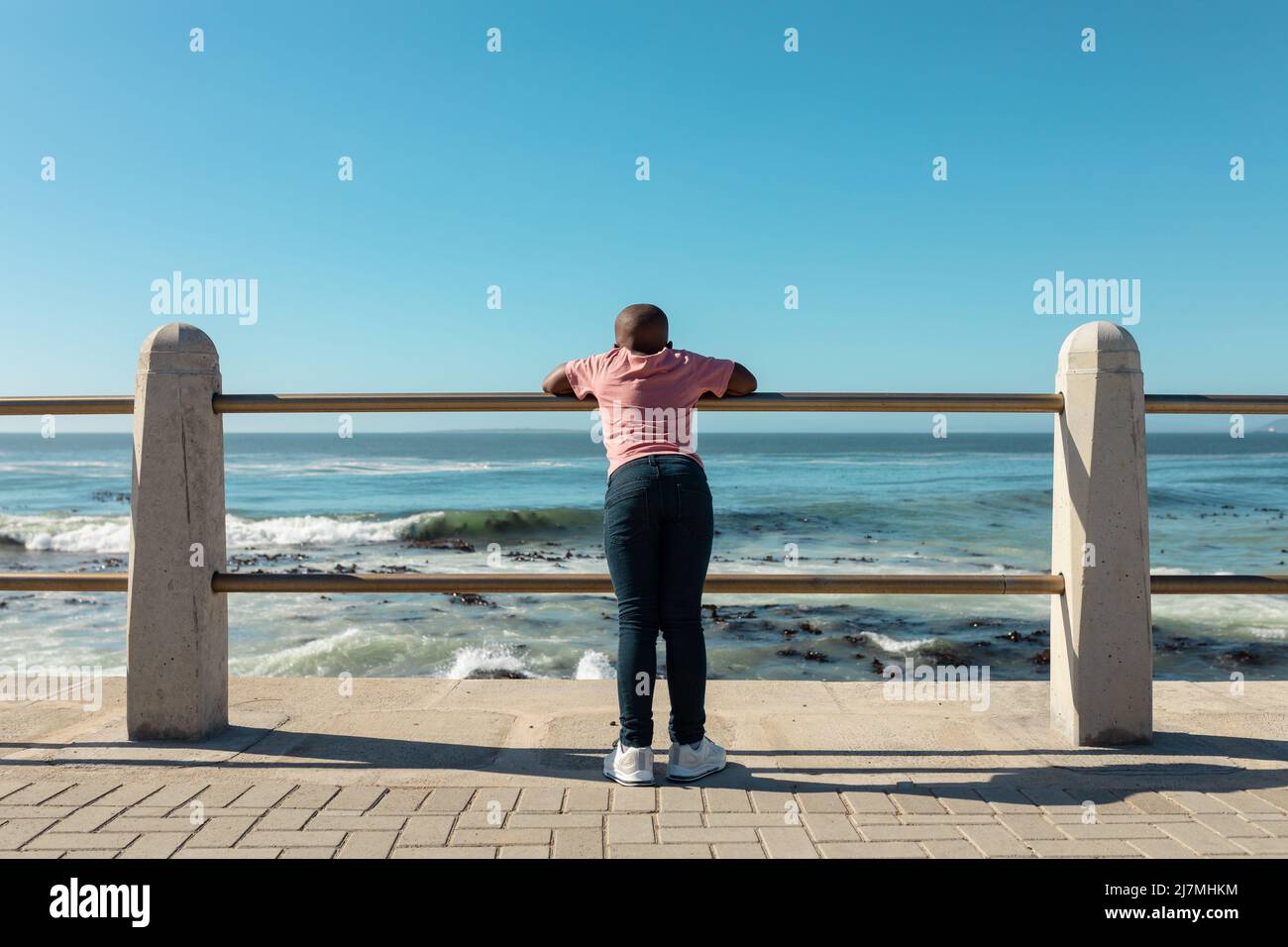 African american boy leaning on railing of promenade looking at sea against blue sky with copy space Stock Photo
