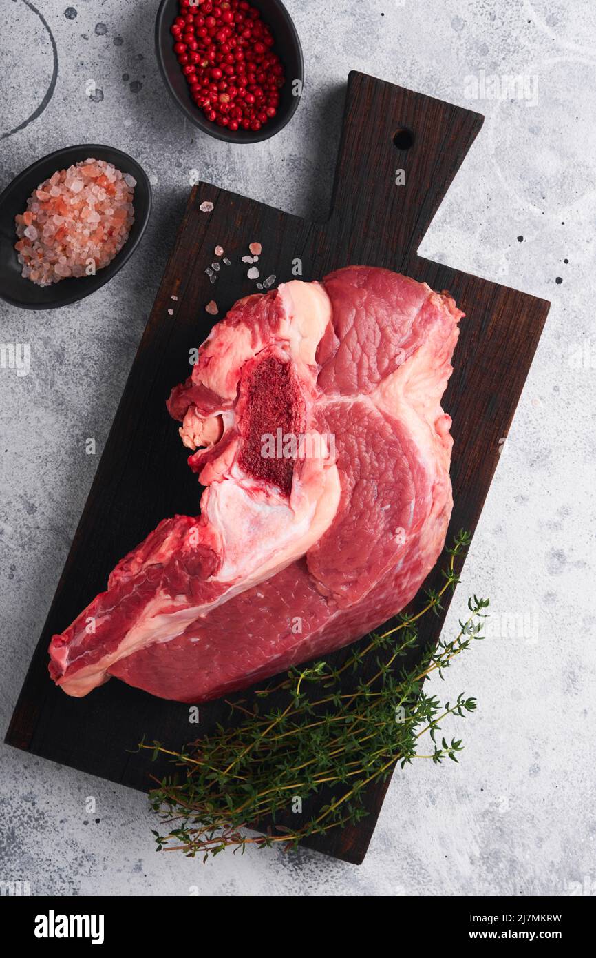 Raw beef meat. Rough piece of meat on bone for roast or soup with salt, pepper, thyme and rosemary on old grey concrete table background. Entrecote. R Stock Photo