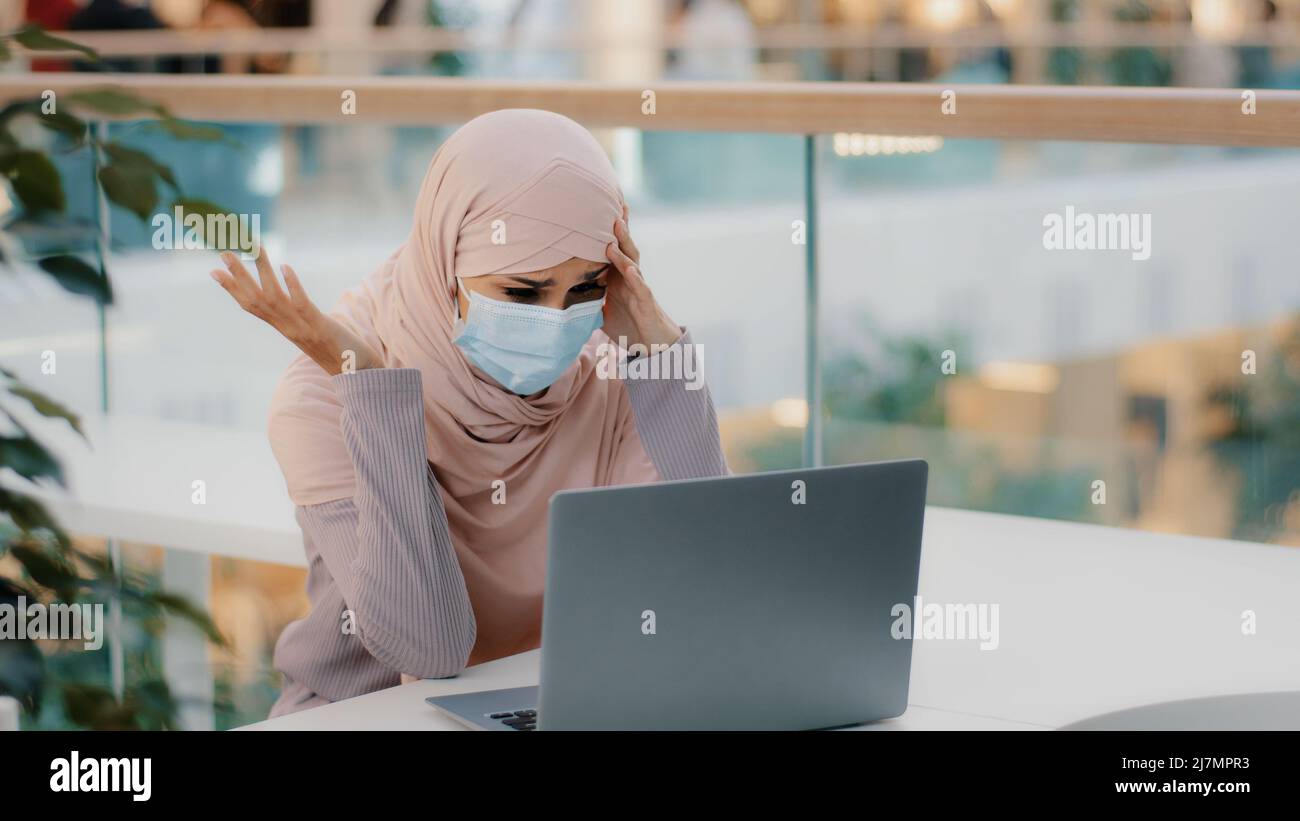 Young arab woman in hijab wearing protective mask reading email on laptop receiving bad news girl upset shocked by notification dismissal from job Stock Photo