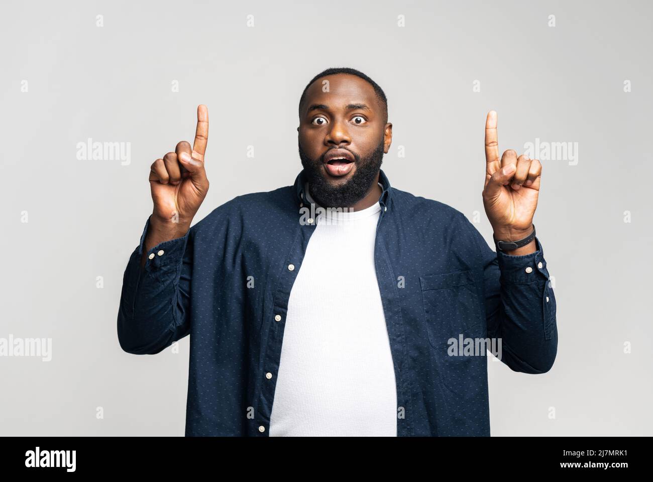 Amused African-American man points index fingers up, looking at the camera with shocked face expression, surprised black guy in casual wear pays attention on promotion, isolated on gray background Stock Photo