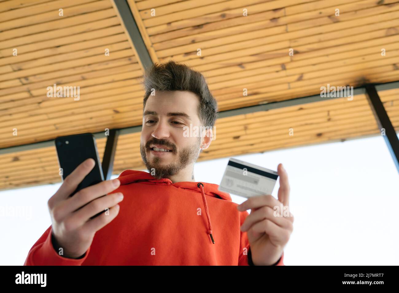 Happy young man looking phone screen and holding credit card for ordering products in internet. Fast online shopping. Stock Photo