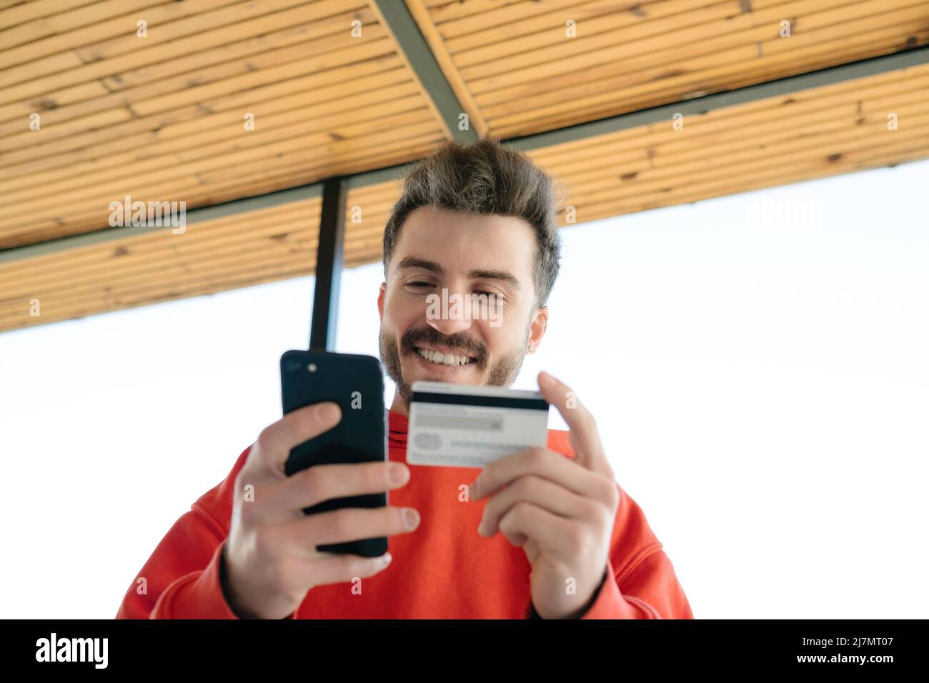 Fast online shopping. Happy young man using phone and credit card for ordering products in internet. Stock Photo