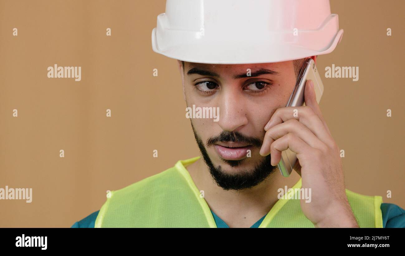 Close up shot of young hispanic man engineer construction foreman builder in special safety helmet talking cell phone with building worker discussing Stock Photo