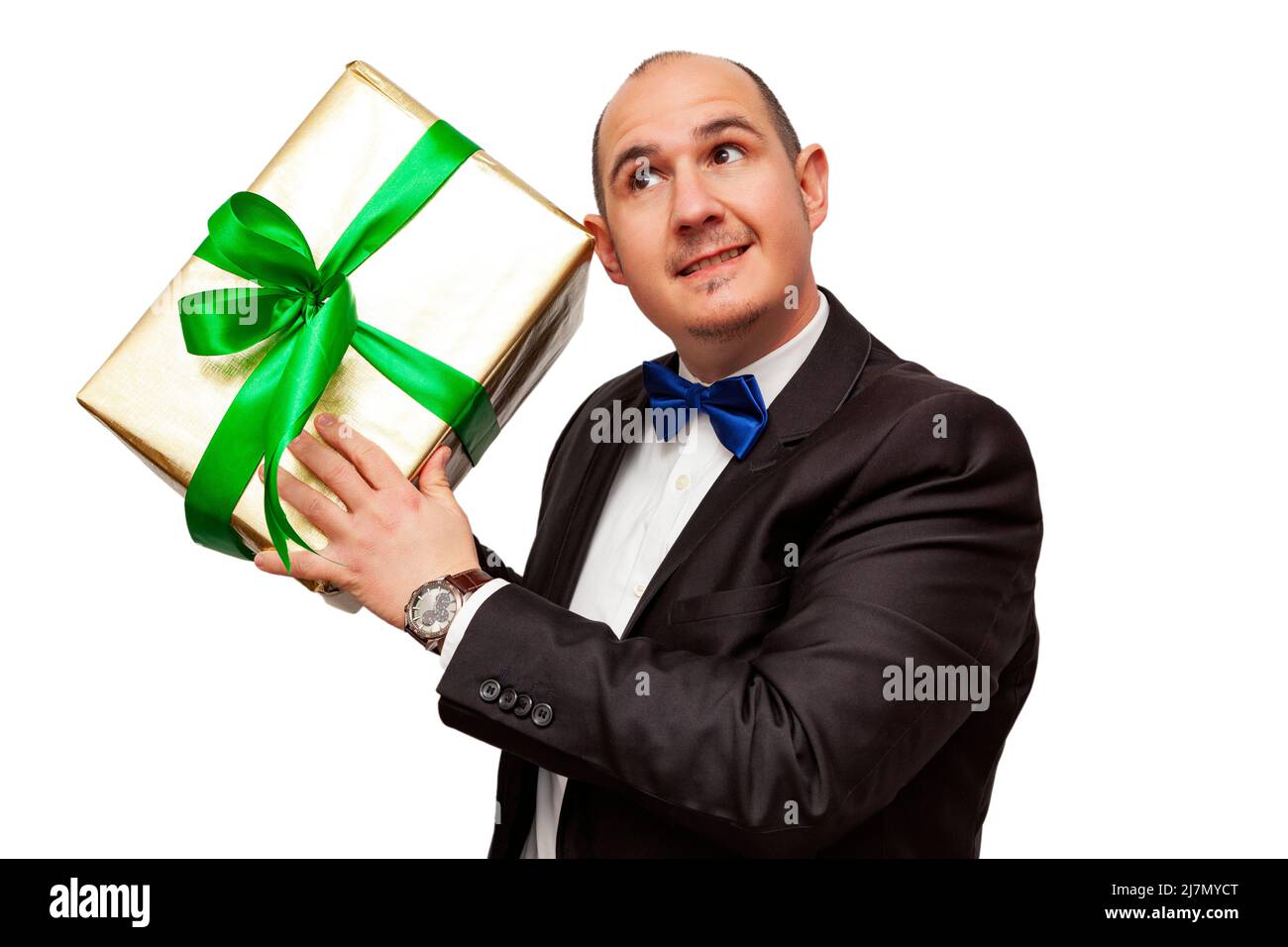 A smiling bald Caucasian adult male wearing a black suit, white shirt and blue bow tie is holding in his hands a gift wrapped with gold foil and a gre Stock Photo