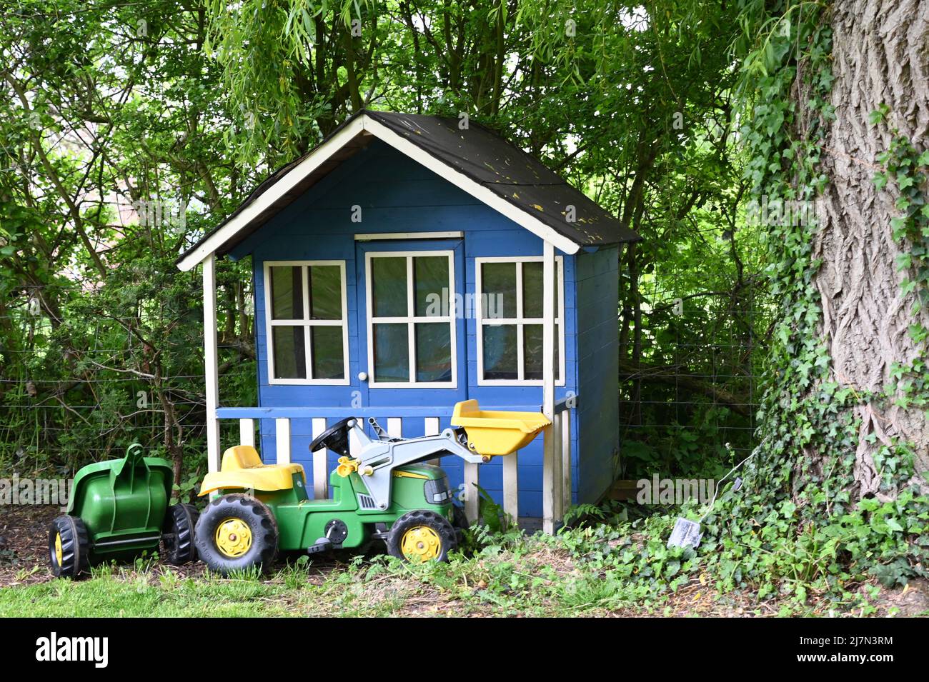 A blue child's wooden play house sitting in front of a hedge by the side of a willow tree with a John Deer child's plastic tractor in front of it Stock Photo