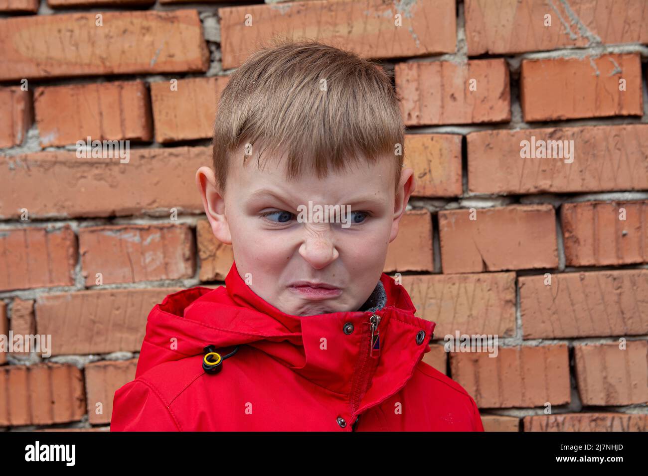 The boy is grimacing in anger. Angry face of a boy in a red jacket. Portrait of a caucasian angry boy. Stock Photo