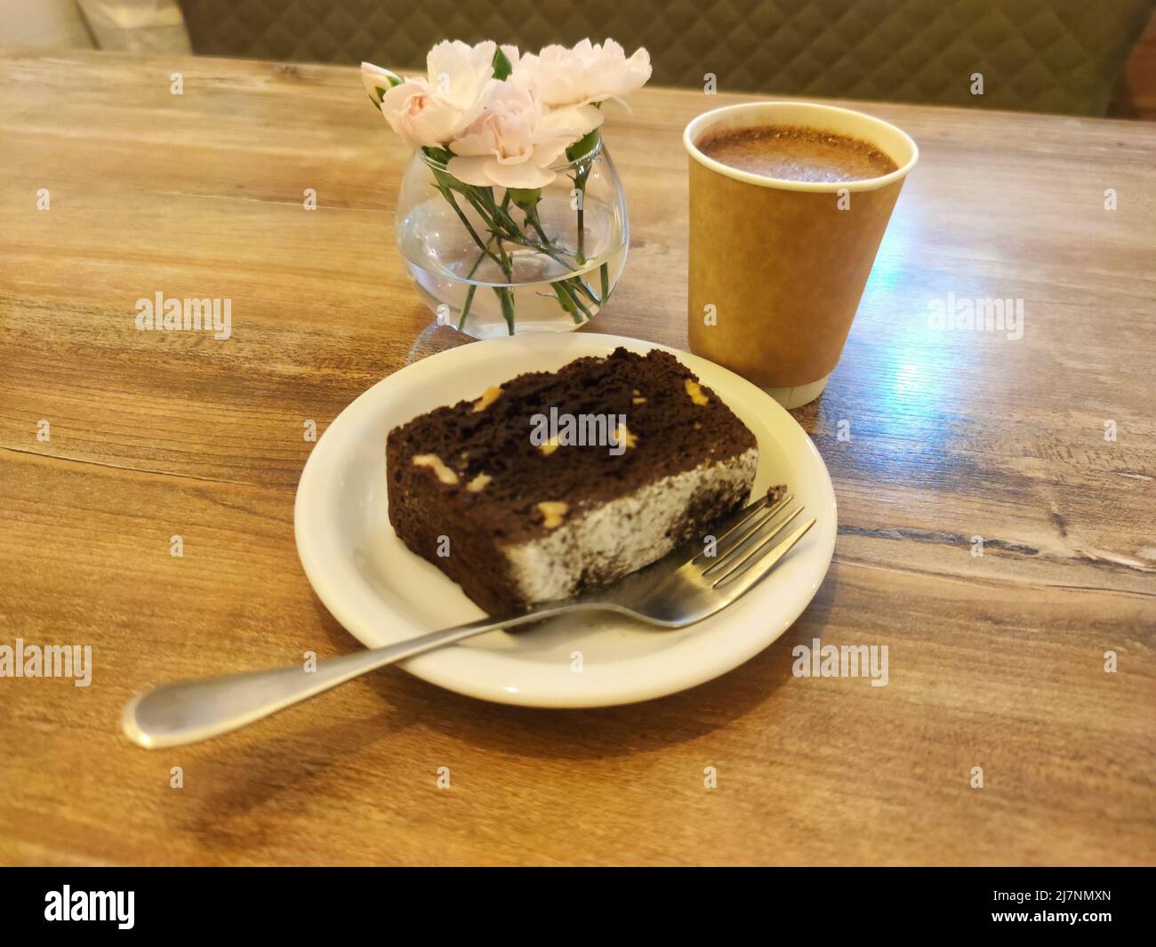 A piece of chocolate brownie cake on the plate, cacao and flowers on the wooden table. Morning coffee, cappuccino, latte and pastry. Pale pink carnati Stock Photo