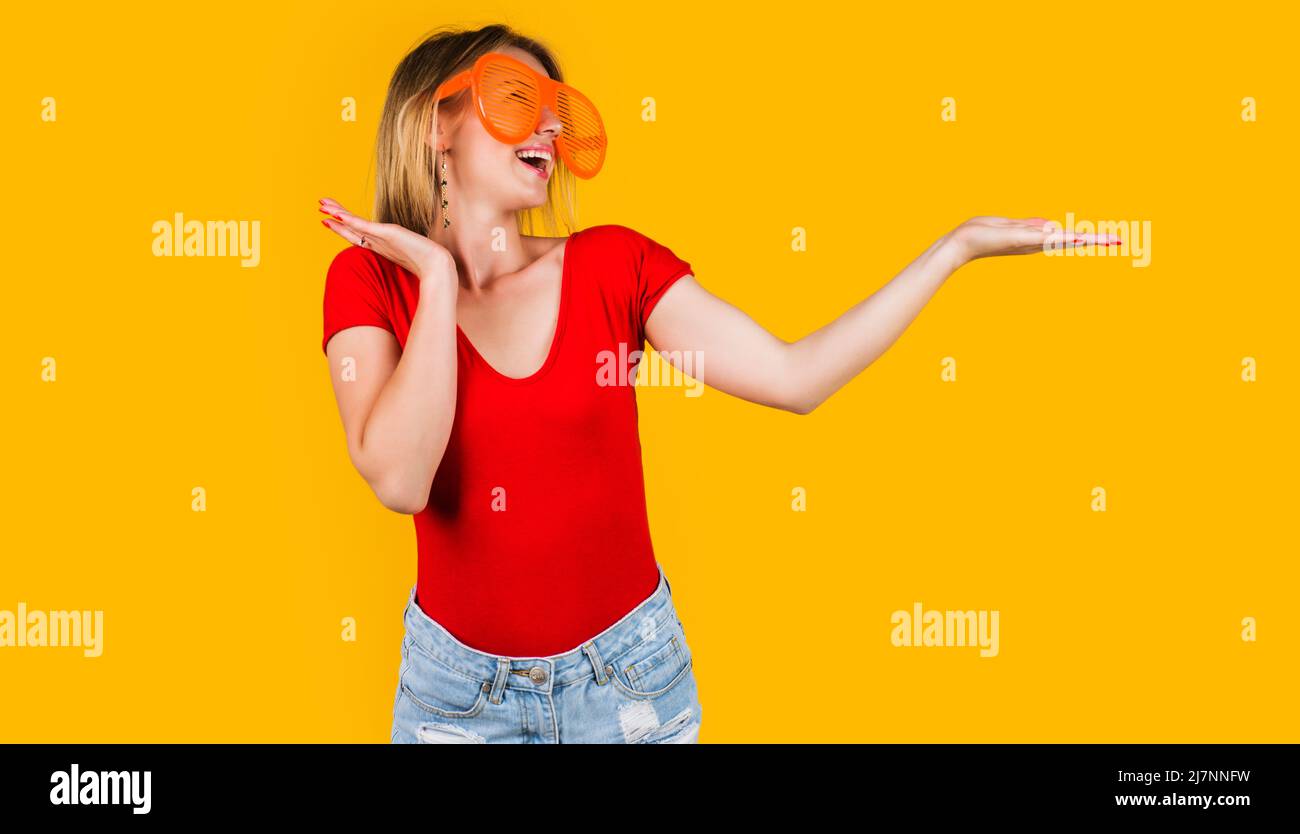 Happy girl in funny glasses presenting product holds something on palm. Woman shows hand gesture. Stock Photo