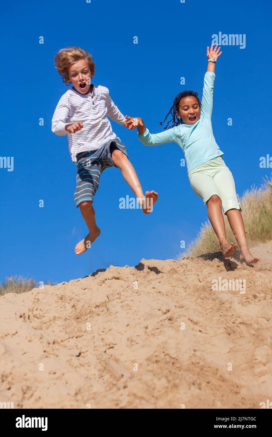 Two children an African American biracial girl and a blonde boy playing jumping and laughing in the dunes of a sandy beach Stock Photo